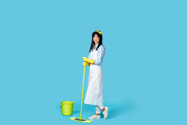 woman in rubber gloves and cleaner apron washing floor with mop on blue background. Full length - Fotó, kép