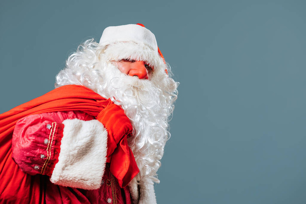 Santa Claus red nose on the blue background front view is leaning under the weight of a red bag with gifts. He holds his burden with his red gloved hands. Looks into the camera. - Fotografie, Obrázek