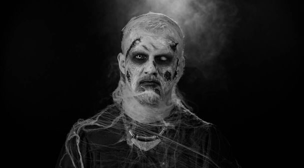 Зловещий человек с ужасным страшным зомби-макияжем на Хэллоуин в костюмах, строящий рожи, смотрящий зловеще на камеру, пытающийся напугать. Мертвый парень с ранеными кровавыми шрамами лица изолированы на черном фоне - Фото, изображение