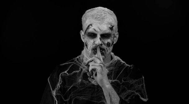 Shh be quiet. Sinister man with horrible scary Halloween zombie makeup in costume presses index finger to lips makes silence gesture sign do not tells secret. Dead guy with wounded bloody scars face - Photo, Image