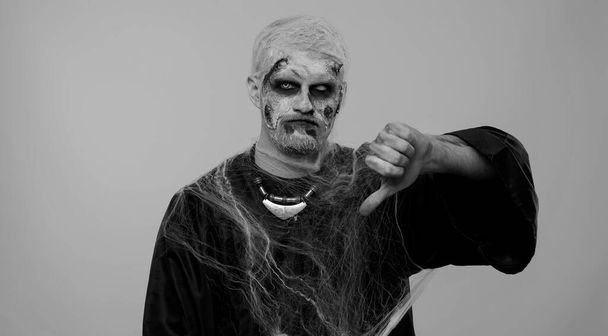 Зловещий человек в костюме сумасшедшего зомби на Хэллоуин с окровавленными ранеными шрамами на лице, показывающими большой палец на знаке жеста, выражающими недовольство, неодобрение, недовольство, неприязнь. Косплей нежити - Фото, изображение