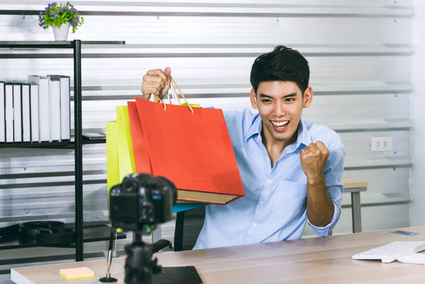 Um jovem asiático influência macho vlogger polegares para cima enquanto segurando sacos de compras em transmissão ao vivo, youtuber conceito - Foto, Imagem