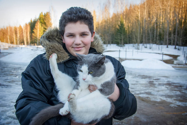 In the spring on the street, a boy holds a large gray-white cat in his arms. - Valokuva, kuva