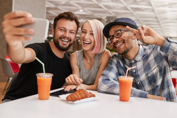 Happy multiracial men and woman in casual clothes smiling and taking selfie via smartphone, while sitting at table with croissant and cups of fresh juice on weekend day in restaurant - Foto, Bild