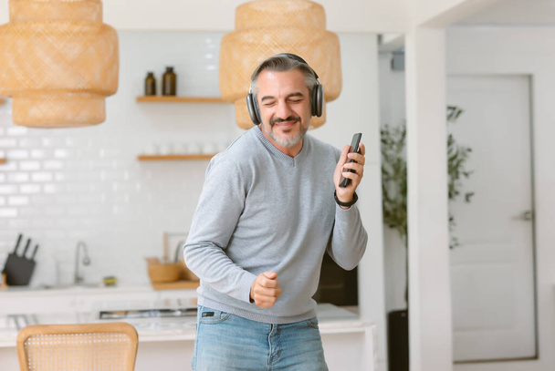 Boldog jóképű középkorú férfi vezeték nélküli fejhallgató hallgatja a zenét okostelefonnal, tánc és mosolygós otthon hangulatos boho belső. Zeneszerető koncepció - Fotó, kép