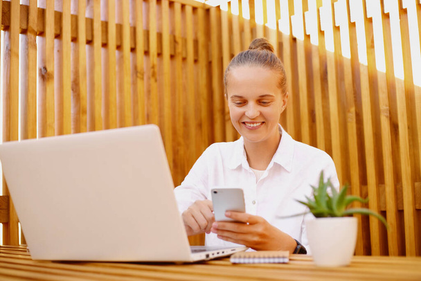 Portrait of smiling positive woman with wearing white shirt working on laptop against wooden wall and using cell phone, expressing good emotions, checking her email. - Valokuva, kuva