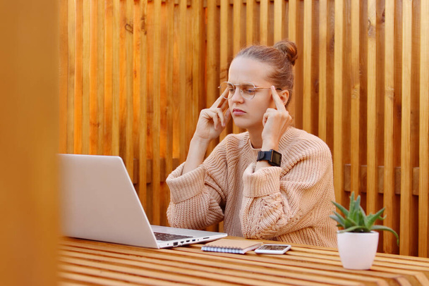 Tired exhausted young adult attractive woman with bun hairstyle wearing beige sweater and glasses working on laptop, feels terrible headache, massaging her temples, sitting against wooden wall. - Valokuva, kuva