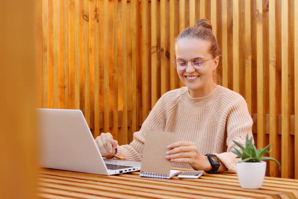 Horizontal shot of positive optimistic pleased woman with bun hairstyle wearing beige jumper and spectacles working on laptop and checking her timetable in paper notebook, posing against wooden wall. - Valokuva, kuva