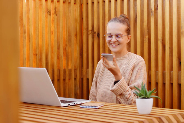 Horizontal shot of winsome smiling woman with bun hairstyle holding smart phone and recording voice message, female wearing beige sweater working on laptop against wooden wall. - Φωτογραφία, εικόνα