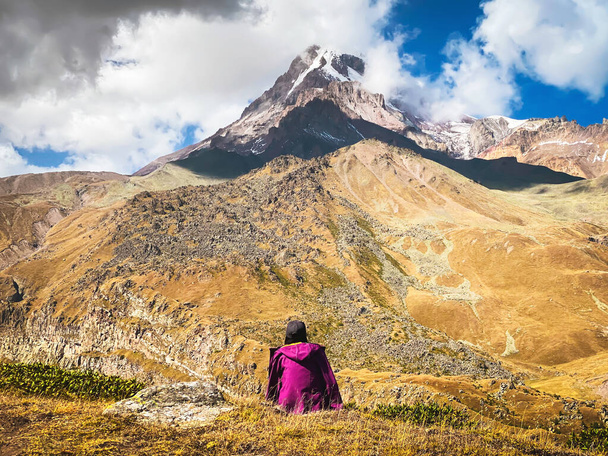 Przemyślana kobieta siedzi na trawie podziwiać piękne góry Kazbek śnieżny szczyt jesienią. Wycieczka na lodowiec Gergeti solo. Podróżowanie na Kaukazie - Zdjęcie, obraz