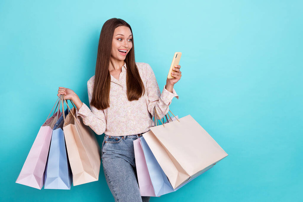 Photo of impressed young woman hold bags look telephone wear white shirt jeans isolated on blue color background. - Foto, afbeelding
