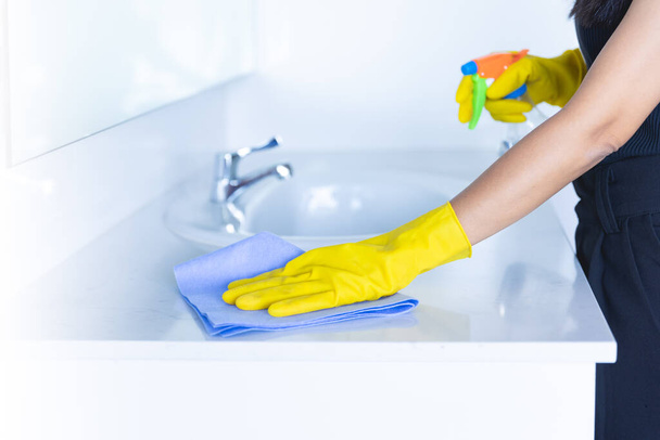 closeup γυναίκες χέρι υπηρέτρια ή Charwoman καθαρισμού σύγχρονη νέα λεκάνη στο μπάνιο. Νερό βρύσης καθαρό χρησιμοποιώντας κίτρινα γάντια και μπλε πανί με ομίχλη ψεκασμού απολυμαντικό. - Φωτογραφία, εικόνα