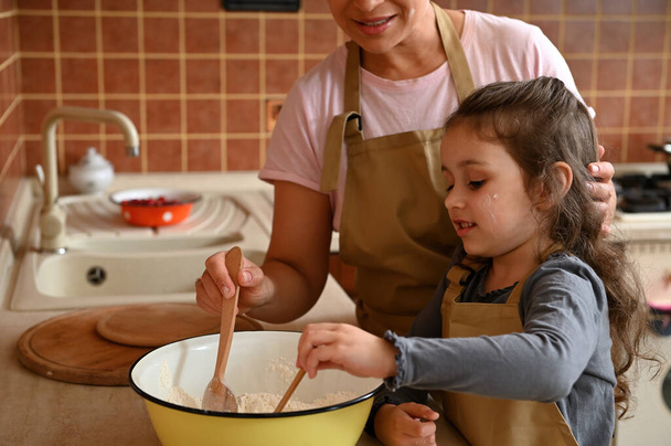Caucasian cute little girl wearing a beige chefs apron, kneading a dough, standing near her mother in the home kitchen and learning preparing pie. Baking concept. Mom and daughter cooking together - Foto, Bild
