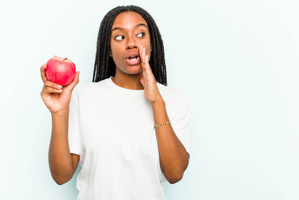 Młoda Afroamerykanka trzymająca jabłko odizolowane na niebieskim tle mówi sekretną wiadomość o gorącym hamowaniu i odwraca wzrok. - Zdjęcie, obraz