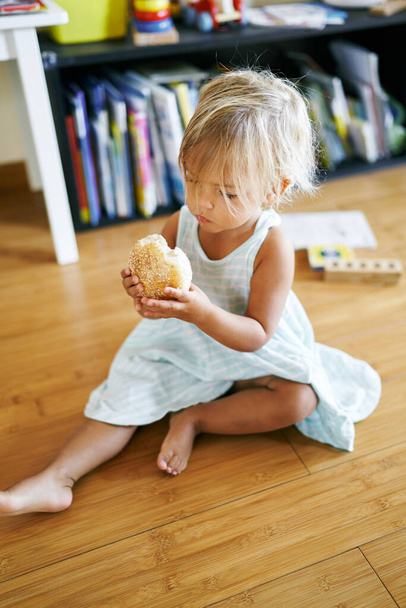 Little girl sits on the floor with a sesame bun in her hands and looks on it. High quality photo - Zdjęcie, obraz