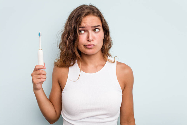Giovane donna caucasica in possesso di spazzolino elettrico isolato sfondo blu confuso, si sente dubbioso e insicuro. - Foto, immagini