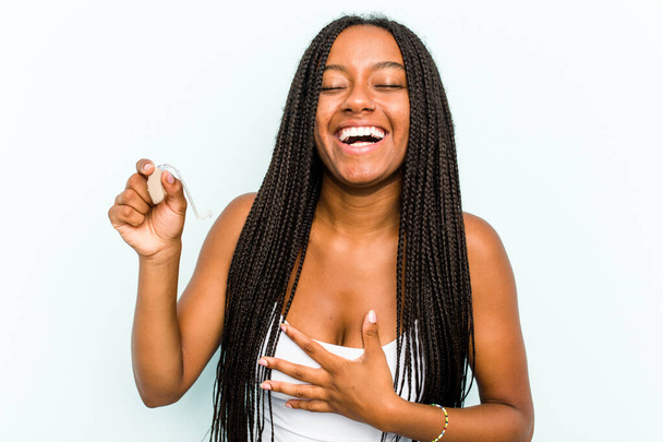Young African American woman holding hearing aid isolated on blue background laughs out loudly keeping hand on chest. - Foto, afbeelding