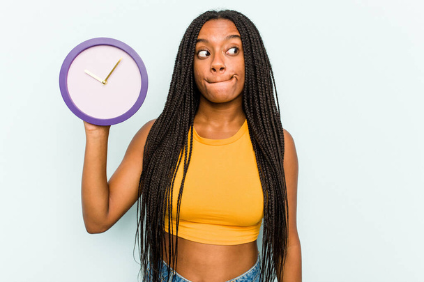 Nuori afroamerikkalainen nainen pitämällä kellon eristetty sinisellä taustalla sekava, tuntuu epäilyttävältä ja epävarma. - Valokuva, kuva