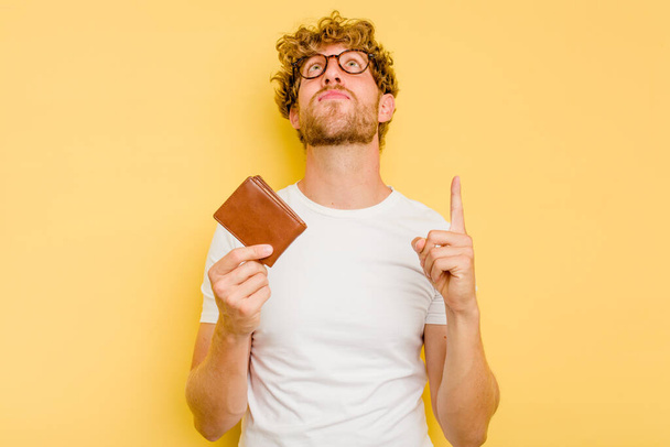 Young caucasian man holding a wallet isolated on yellow background pointing upside with opened mouth. - Photo, image