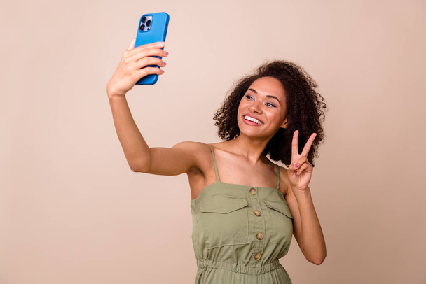 Photo of nice pretty gorgeous girl with wavy hairdo dressed khaki overall making selfie show v-sign isolated on beige color background. - Fotó, kép