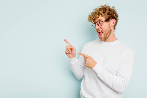 Young caucasian man isolated on blue background points with thumb finger away, laughing and carefree. - Foto, afbeelding