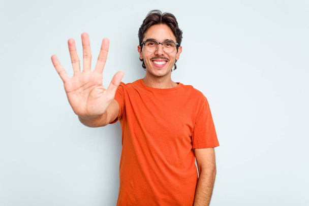 Νεαρός Ισπανόφωνος απομονωμένος σε μπλε φόντο χαμογελώντας χαρούμενος δείχνοντας το νούμερο πέντε με τα δάχτυλα. - Φωτογραφία, εικόνα