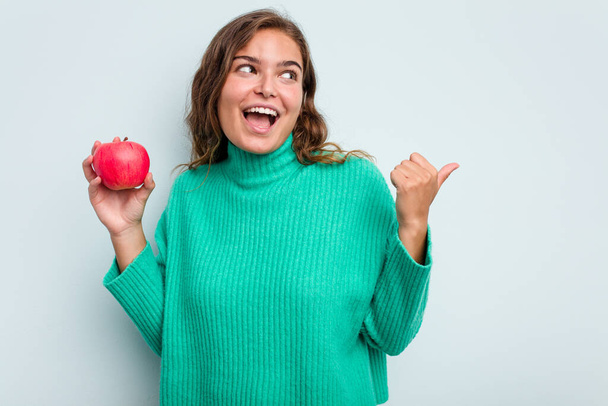 Young caucasian woman with an apple isolated on blue background points with thumb finger away, laughing and carefree. - Valokuva, kuva