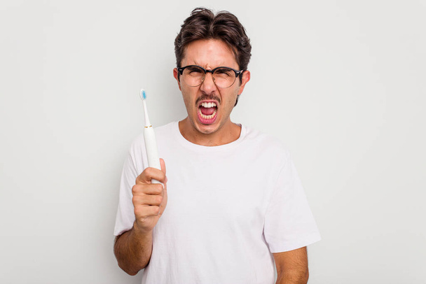 Νεαρός Ισπανός που κρατάει την ηλεκτρική οδοντόβουρτσα απομονωμένη σε λευκό φόντο ουρλιάζοντας πολύ θυμωμένος και επιθετικός. - Φωτογραφία, εικόνα