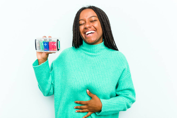 Молода афроамериканська жінка, яка тримає акумуляторну коробку на синьому фоні, сміється з того, що тримає руки за груди.. - Фото, зображення
