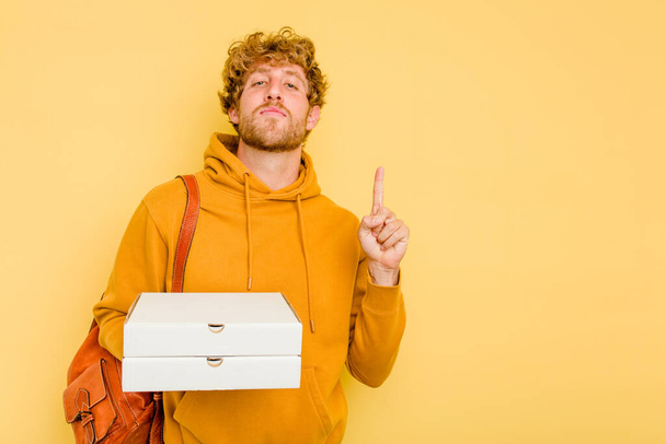 Fiatal diák férfi gazdaság pizza elszigetelt sárga háttér mutatja az első számú ujját. - Fotó, kép