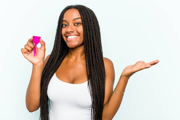 Young African American woman holding menstrual cup isolated on blue background showing a copy space on a palm and holding another hand on waist. - Foto, afbeelding