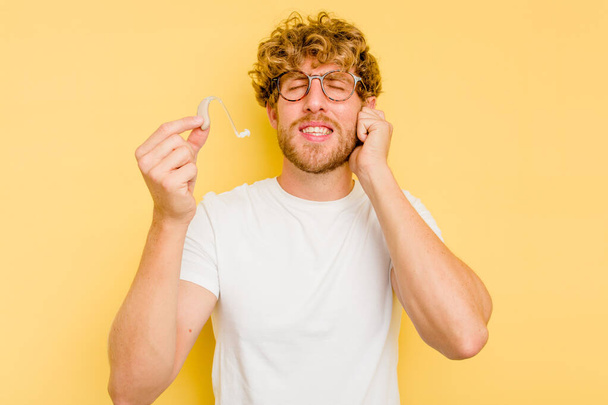 Jonge blanke man met gehoorapparaat geïsoleerd op gele achtergrond die oren met handen bedekt. - Foto, afbeelding