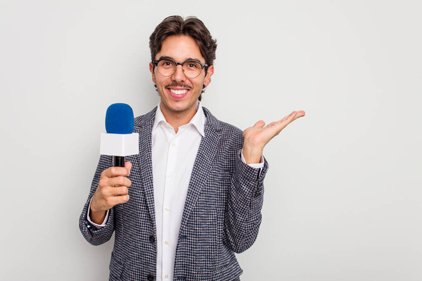 Young hispanic TV presenter man isolated on white background showing a copy space on a palm and holding another hand on waist. - 写真・画像