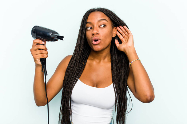 Νεαρή Αφρο-Αμερικανίδα που κρατάει ένα στεγνωτήρα μαλλιών απομονωμένο σε μπλε φόντο προσπαθώντας να ακούσει ένα κουτσομπολιό. - Φωτογραφία, εικόνα