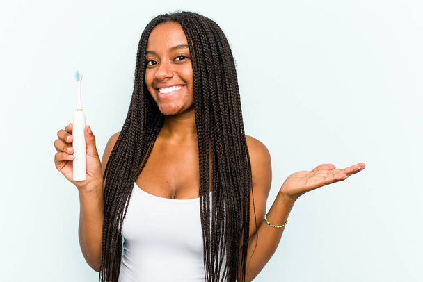 Jeune femme afro-américaine tenant une brosse à dents électrique isolée sur fond bleu montrant un espace de copie sur une paume et tenant une autre main à la taille. - Photo, image