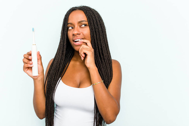 Jonge Afro-Amerikaanse vrouw met elektrische tandenborstel geïsoleerd op blauwe achtergrond ontspannen denken over iets op zoek naar een kopieerruimte. - Foto, afbeelding