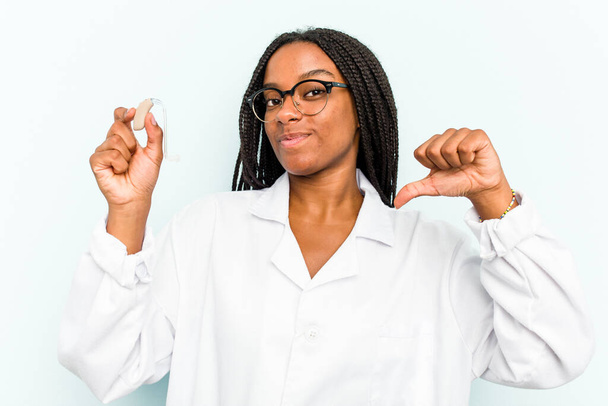 La joven otorrinolaringóloga afroamericana que sostiene un audífono aislado sobre fondo azul se siente orgullosa y segura de sí misma, ejemplo a seguir. - Foto, imagen