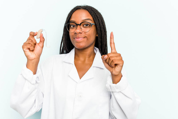Giovane donna otorinolaringoiatra afroamericana con un apparecchio acustico isolato su sfondo blu che mostra il numero uno con il dito. - Foto, immagini