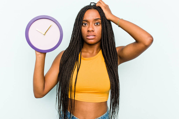 Молодая афроамериканка, держащая часы, изолированные на синем фоне, была шокирована, она вспомнила важную встречу. - Фото, изображение