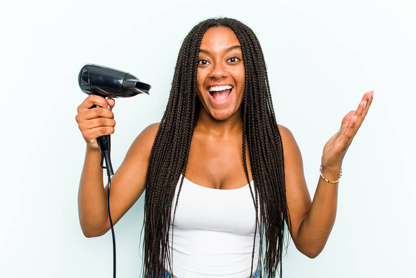 Молода афроамериканська жінка з перукарем, ізольованою на синьому фоні, отримує приємний сюрприз, захоплені і підняті руки.. - Фото, зображення