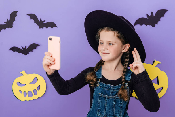 Smiling little girl kid in big hat holding telephone shooting making funky selfies showing v-sign symbol, isolated on purple color background in studio decorated yellow paper pumpkins and black bats - Фото, изображение