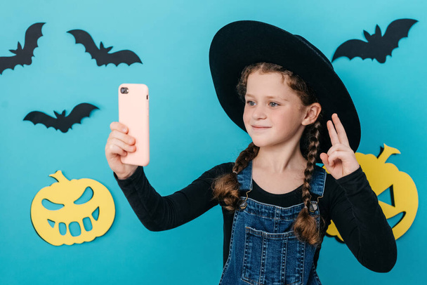 Beautiful little girl kid in black hat holding telephone shooting making funky selfies showing v-sign symbol, isolated on blue color background in studio decorated yellow paper pumpkins and small bats - Foto, imagen