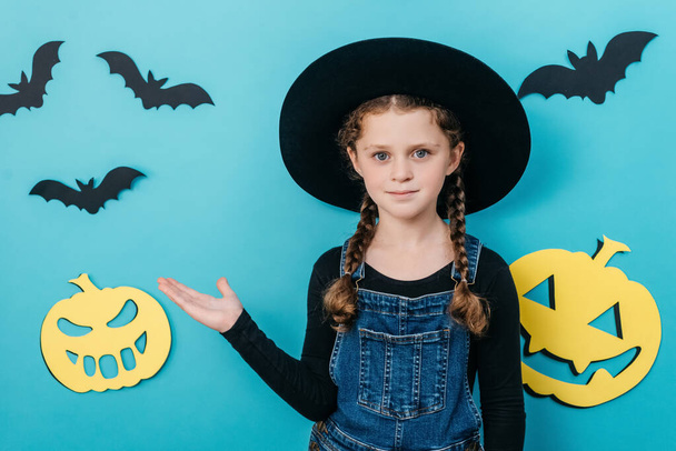Portrait of little girl kid in hat point palm aside on workspace copy space mockup promo area, posing isolated over blue color background wall in studio decorated paper pumpkins and bats. Halloween - Φωτογραφία, εικόνα