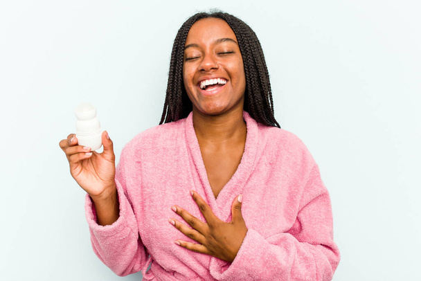 Young African American woman wearing bathrobe holding a deodorant isolated on blue background laughs out loudly keeping hand on chest. - Valokuva, kuva