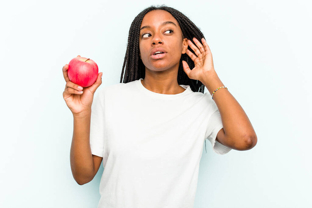 Młoda Afroamerykanka trzymająca jabłko na niebieskim tle próbująca słuchać plotek. - Zdjęcie, obraz
