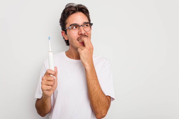 Νεαρός Ισπανόφωνος κρατώντας την ηλεκτρική οδοντόβουρτσα απομονωμένη σε λευκό φόντο χαλαρός σκεπτόμενος κάτι κοιτάζοντας ένα χώρο αντιγραφής. - Φωτογραφία, εικόνα