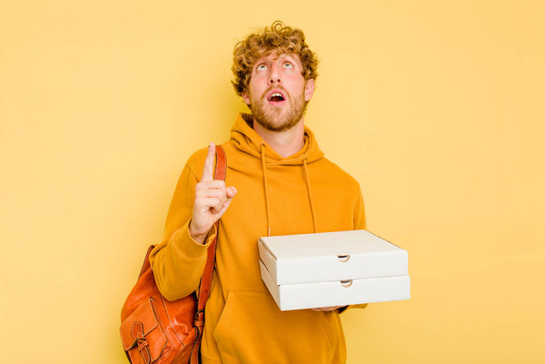 Junger Student hält Pizzen isoliert auf gelbem Hintergrund und zeigt mit geöffnetem Mund auf den Kopf. - Foto, Bild