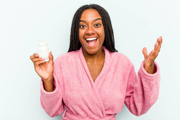 Молодая афроамериканка в халате с дезодорантом на синем фоне получает приятный сюрприз, возбужденную и поднимающую руки. - Фото, изображение