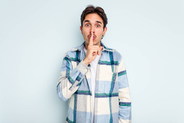 Young hispanic man isolated on blue background keeping a secret or asking for silence. - Foto, imagen