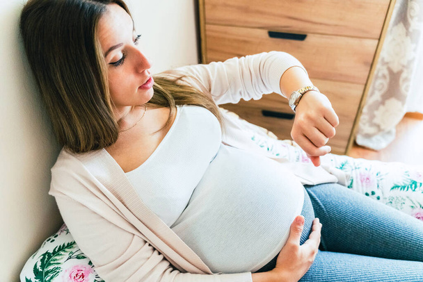 El tiempo de embarazo va al hospital. Embarazada sosteniendo el vientre del bebé, mujer mirando el reloj. Hora del parto, contracciones dolor. Concepto maternidad, embarazo, parto - Foto, Imagen